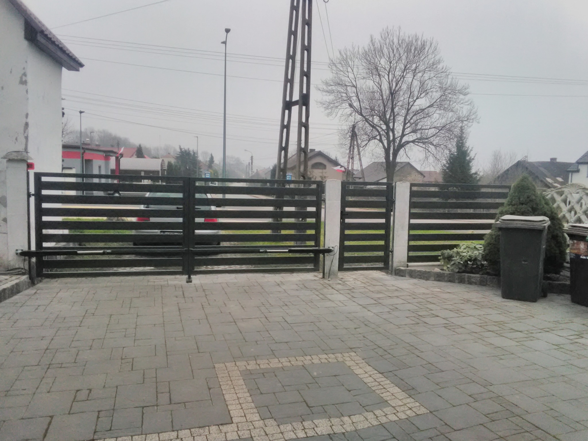 Nowoczesna, pozioma brama wjazdowa z automatyką, widoczna z zewnątrz posesji na tle szarej, pochmurnej pogody, z kostką brukową na podjeździe i słupem energetycznym w tle.