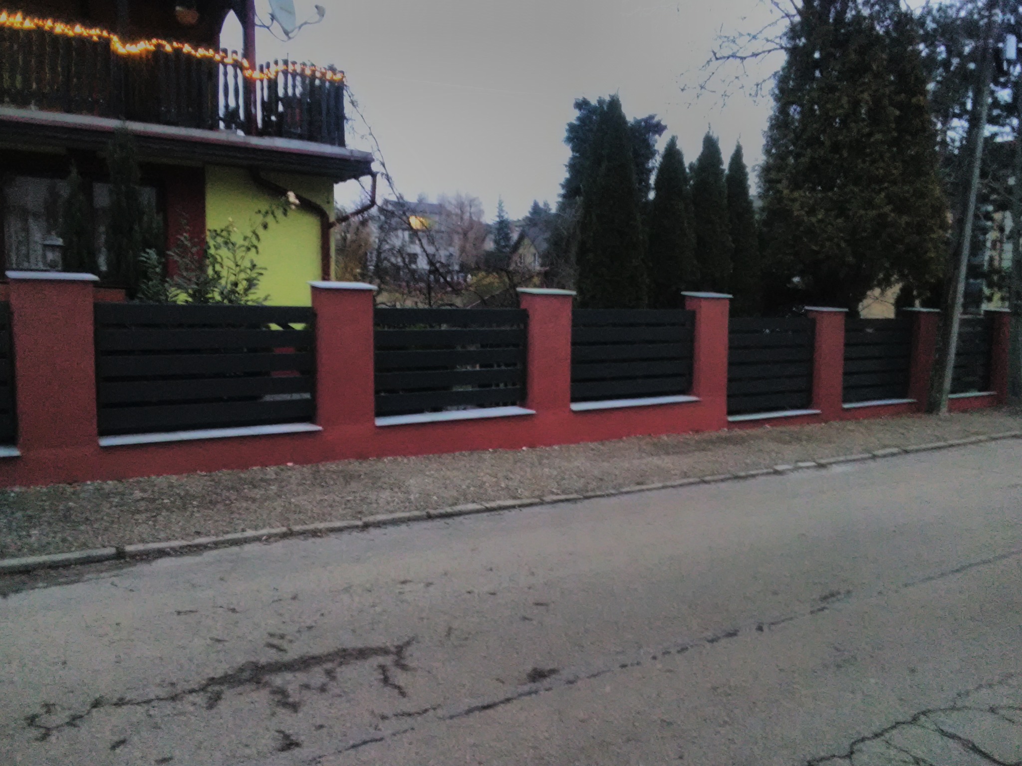 Nowoczesne ogrodzenie z poziomych, ciemnych desek i czerwonych, murowanych słupków, oddzielające posesję od ulicy. W tle widoczny dom z balkonem ozdobionym lampkami.