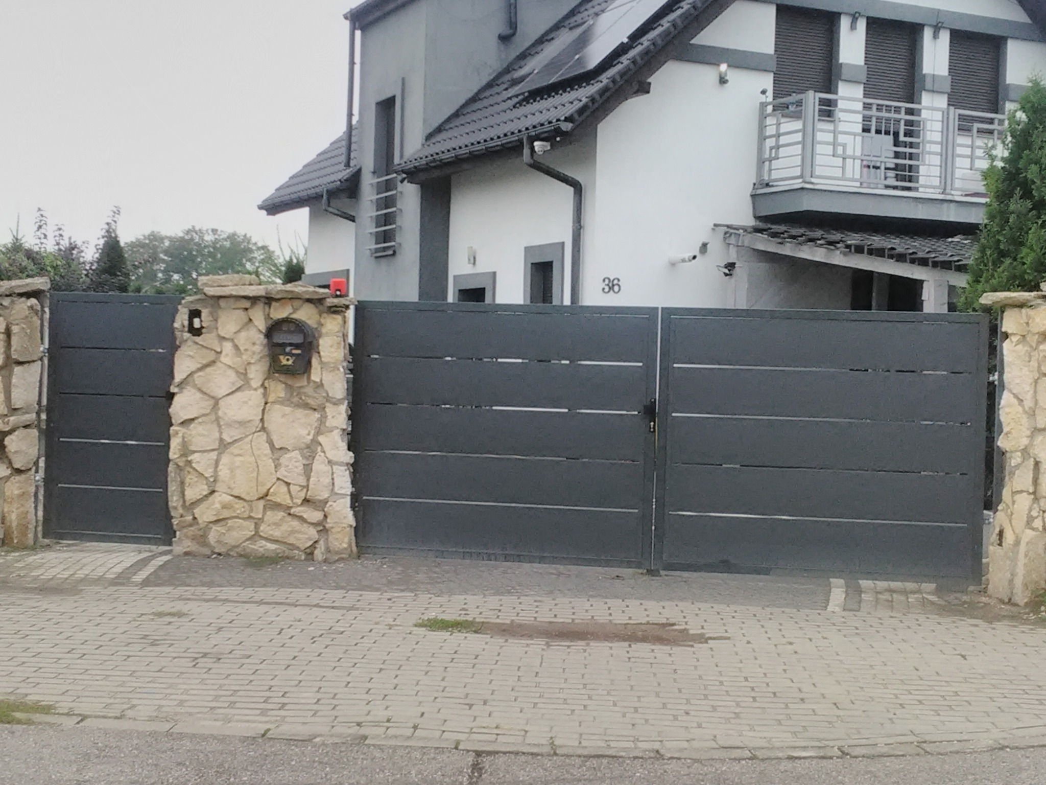 Nowoczesna, pozioma, grafitowa brama wjazdowa z kamiennymi słupkami i skrzynką na listy, na tle domu z balkonem i panelami słonecznymi.