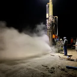 Nocne wiercenie studni głębinowej z użyciem oświetlonej wiertnicy, dwóch pracowników w odzieży roboczej i obłokiem pary wodnej wydobywającej się z otworu.