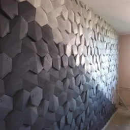 Ściana ozdobiona trójwymiarowymi panelami w kształcie ściętych sześcianów, tworzących gradient od ciemnoszarego do jasnoszarego, z widoczną fakturą betonu i delikatnym oświetleniem padającym z góry.