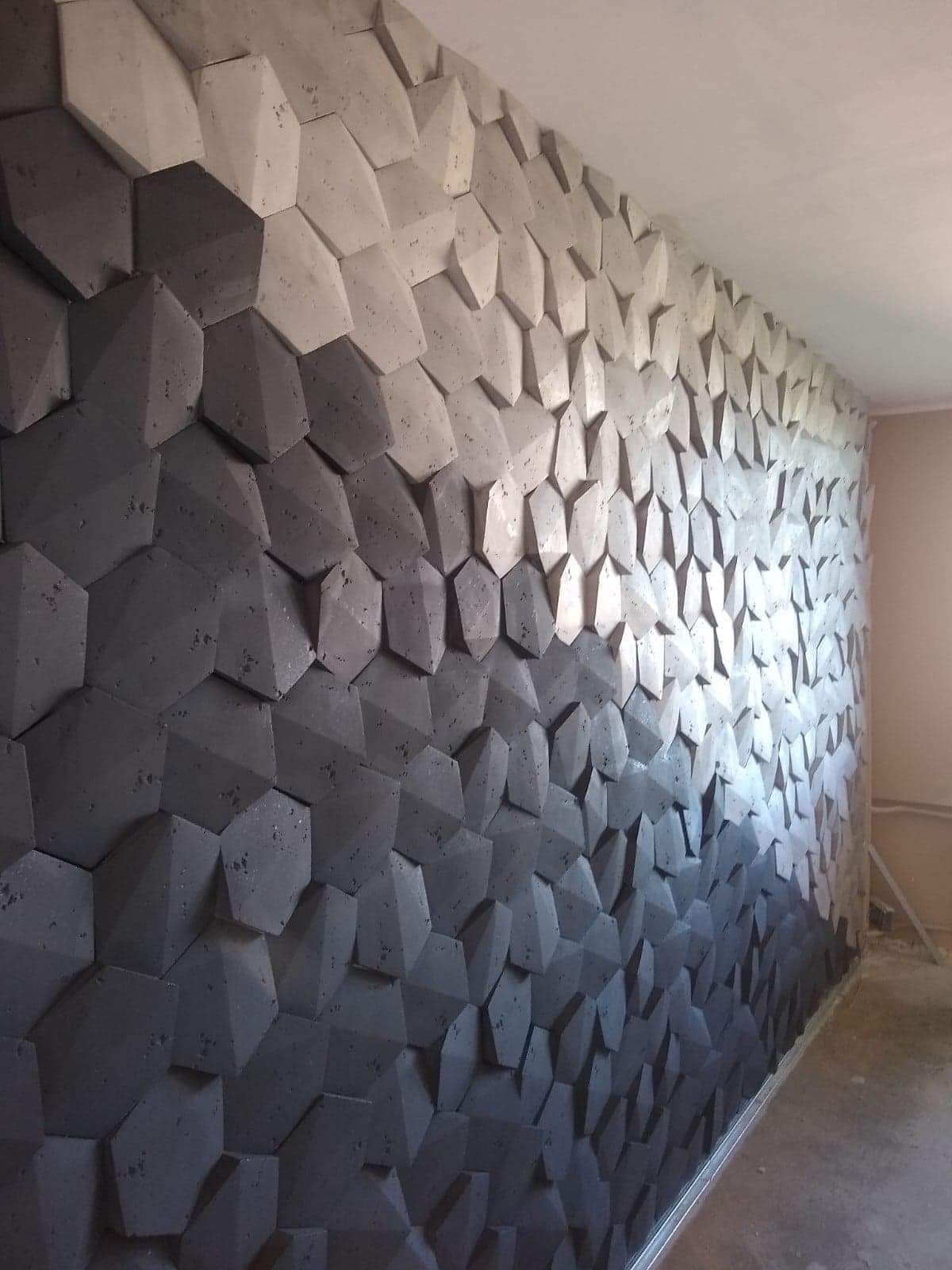 Ściana ozdobiona trójwymiarowymi panelami w kształcie ściętych sześcianów, tworzących gradient od ciemnoszarego do jasnoszarego, z widoczną fakturą betonu i delikatnym oświetleniem padającym z góry.