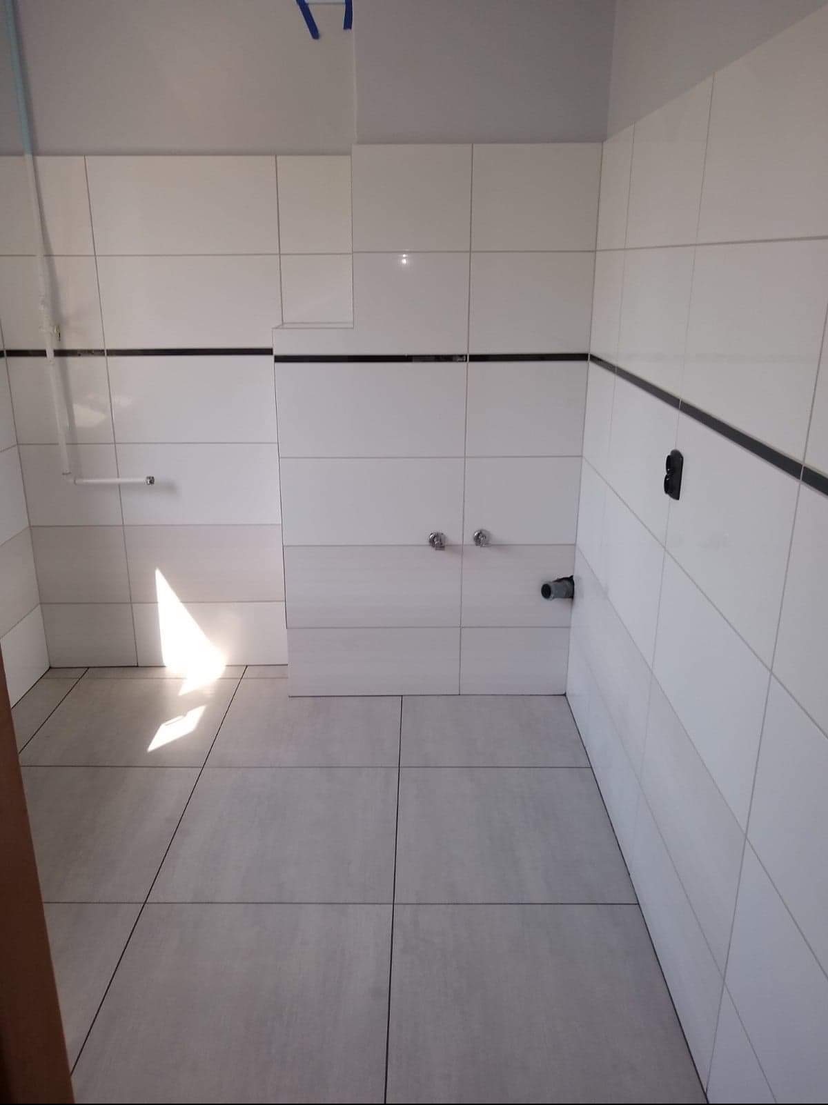 Wykończona łazienka z białymi płytkami ułożonymi w prostokątnym wzorze, ciemnoszary pasek akcentujący wzdłuż górnej krawędzi płytek, dwa zawory na ścianie i rura odpływowa, widoczne niedoskonałości...