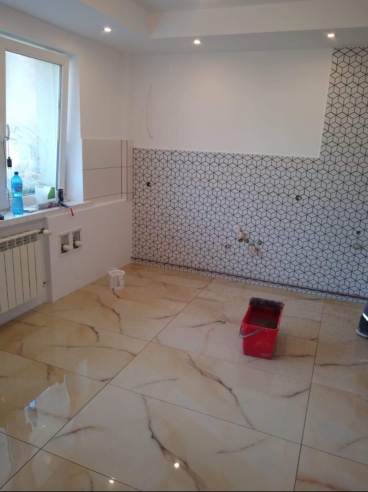 Wnętrze łazienki w trakcie prac: ułożone płytki podłogowe imitujące marmur, częściowo wykończona ściana z geometrycznym wzorem, widoczne instalacje wodne, czerwone wiadro z narzędziami.