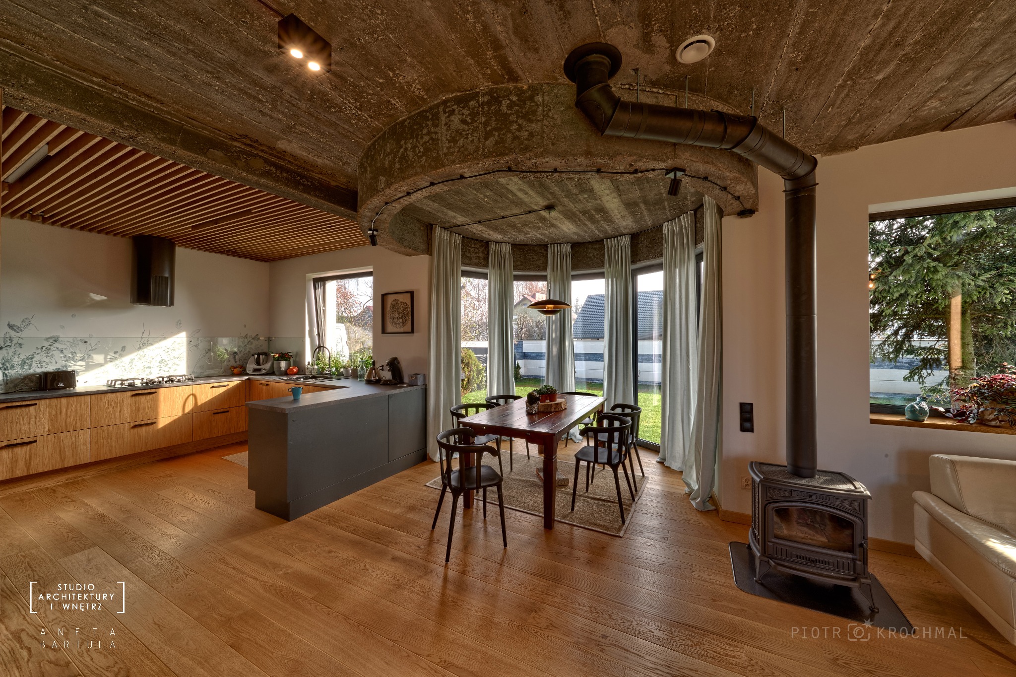 Otwarta przestrzeń mieszkalna z kuchnią i jadalnią, surowy betonowy sufit z okrągłą konstrukcją nad stołem, piec wolnostojący z rurą dymową, drewniana podłoga i duże okna z widokiem na ogród.