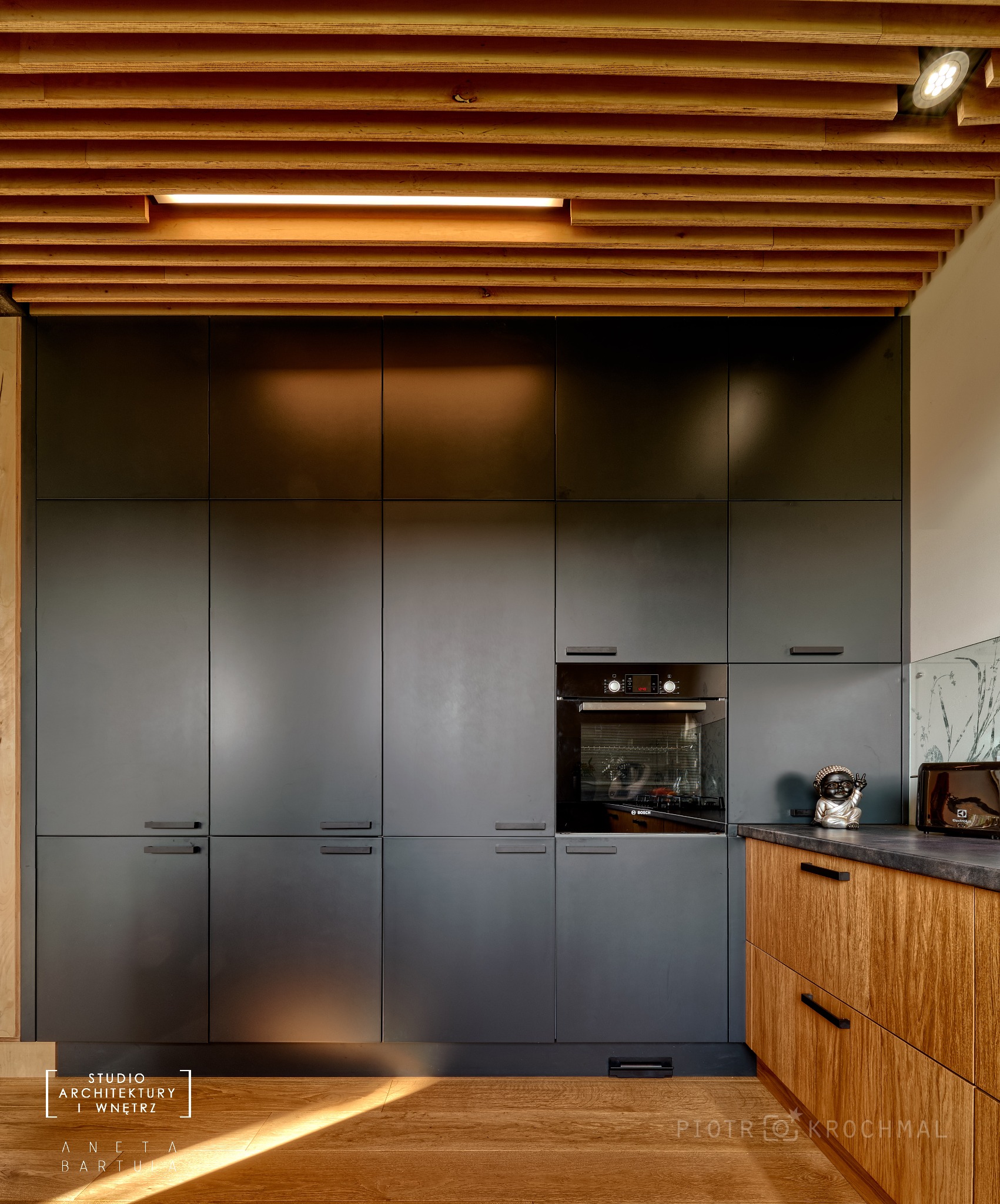 Nowoczesna kuchnia z ciemnymi, matowymi szafkami, drewnianym blatem i sufitem z drewnianych listew z oświetleniem LED, widoczny piekarnik Bosch i dekoracyjna figurka.