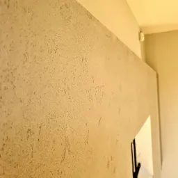 Ściana pokryta beżową, chropowatą fakturą, widoczna z boku, z fragmentem białego narożnika i wystającym elementem w dolnym rogu, oświetlona ciepłym światłem.