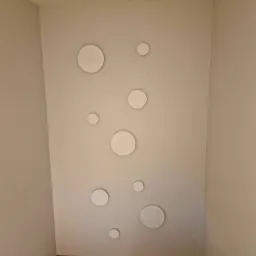 Ściana w rogu pokoju pomalowana na jasny kolor z dekoracyjnymi, okrągłymi elementami o różnych rozmiarach, rozmieszczonymi asymetrycznie. Na podłodze widoczne panele w kolorze drewna.