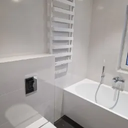 Nowoczesna łazienka w jasnych odcieniach szarości z białą wanną prostokątną, wiszącą toaletą i grzejnikiem drabinkowym, podkreślona ciemną podłogą.