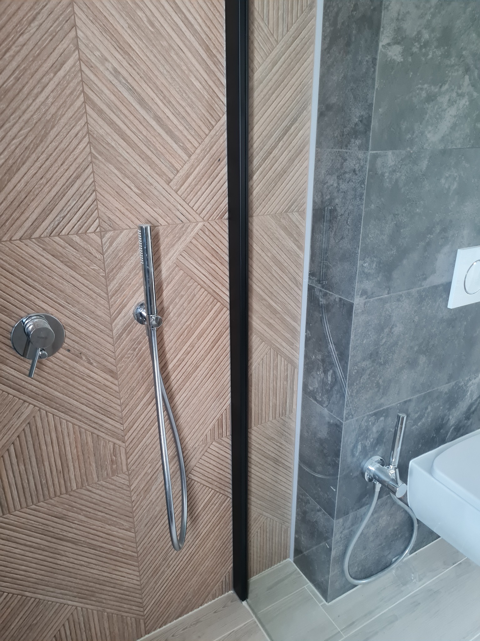 Nowoczesna łazienka z drewnopodobnymi płytkami w strefie prysznica i szarymi płytkami imitującymi kamień w strefie toalety, widoczna armatura i fragment toalety.