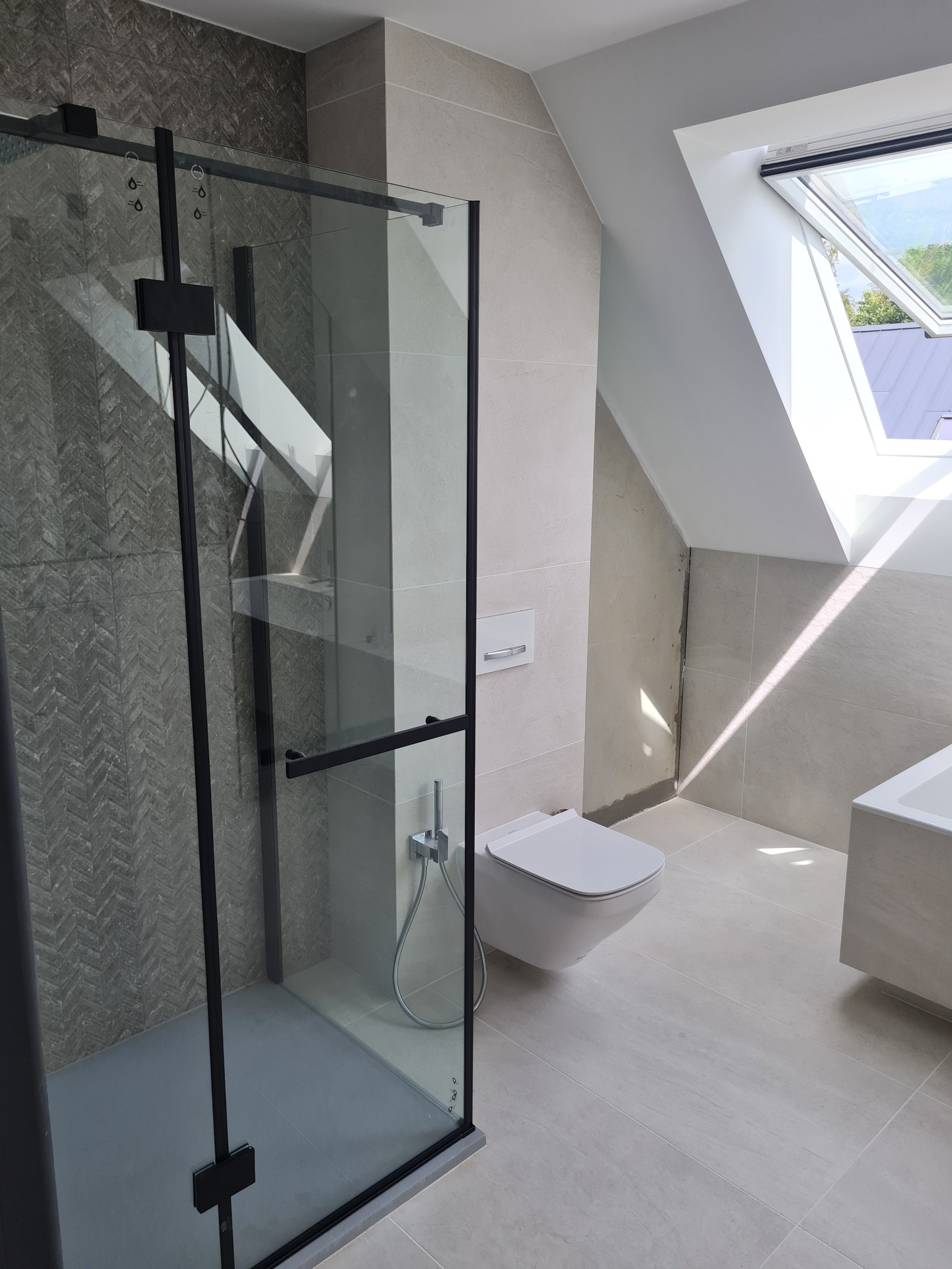 Nowoczesna łazienka na poddaszu z kabiną prysznicową z czarnymi profilami, podwieszaną toaletą i oknem dachowym wpuszczającym jasne światło.