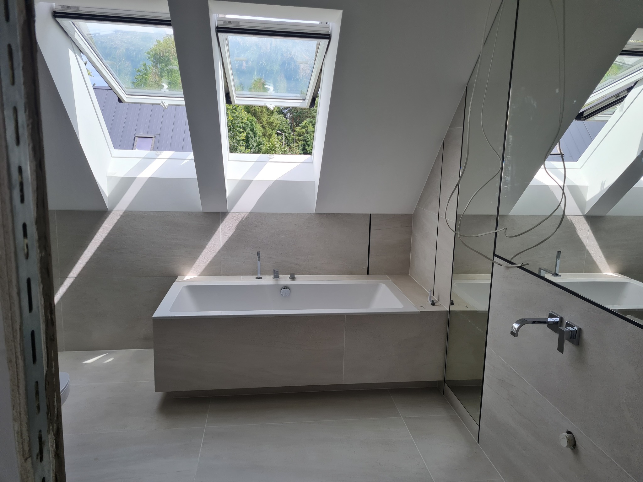 Nowoczesna łazienka na poddaszu z wanną obudowaną płytkami w odcieniach szarości, dwa okna dachowe wpuszczają jasne światło, widok na zieleń za oknem, minimalistyczna armatura.