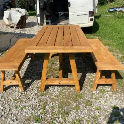 Nowy, drewniany stół piknikowy z dwiema ławkami, pomalowany bejcą na ciepły, miodowy odcień, stoi na żwirowym podłożu, z otwartym bagażnikiem białego vana w tle.