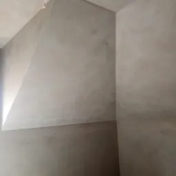 Wnętrze po nałożeniu tynku maszynowego, widoczne nierówności i faktura na ścianach oraz wystający przewód elektryczny z sufitu.