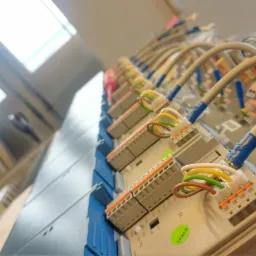 Rząd szarych modułów sterujących z kolorowymi kablami podłączonymi do pomarańczowych złączy, zamontowanych na niebieskiej szynie DIN, widok z dołu pod kątem, z widocznym oknem w tle.