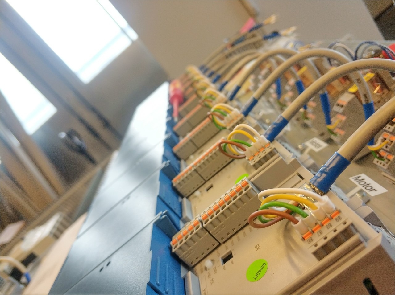 Rząd szarych modułów sterujących z kolorowymi kablami podłączonymi do pomarańczowych złączy, zamontowanych na niebieskiej szynie DIN, widok z dołu pod kątem, z widocznym oknem w tle.