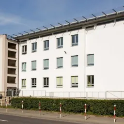 Dwupiętrowy budynek z białymi ścianami i rzędem okien, z zewnętrznymi schodami po prawej stronie i przeszklonym zadaszeniem, widoczny z poziomu ulicy w słoneczny dzień.