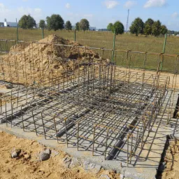 Zbrojenie fundamentu z siatki stalowej na betonowej podstawie, obok pryzma piasku, w tle zielone drzewa i błękitne niebo.