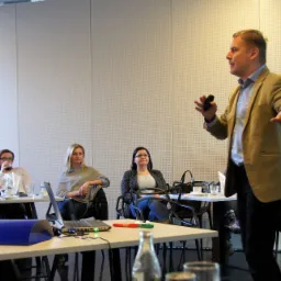 Prelegent w beżowej marynarce gestykuluje podczas prezentacji dla grupy słuchaczy siedzących przy stolikach z laptopami i notatkami w jasnej sali konferencyjnej z białą ścianą w tle.