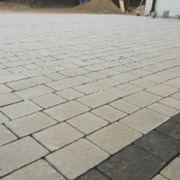 Ułożona jasnoszara kostka brukowa z ciemniejszym pasem obrzeża, widoczny stos kostki i narzędzia w tle.