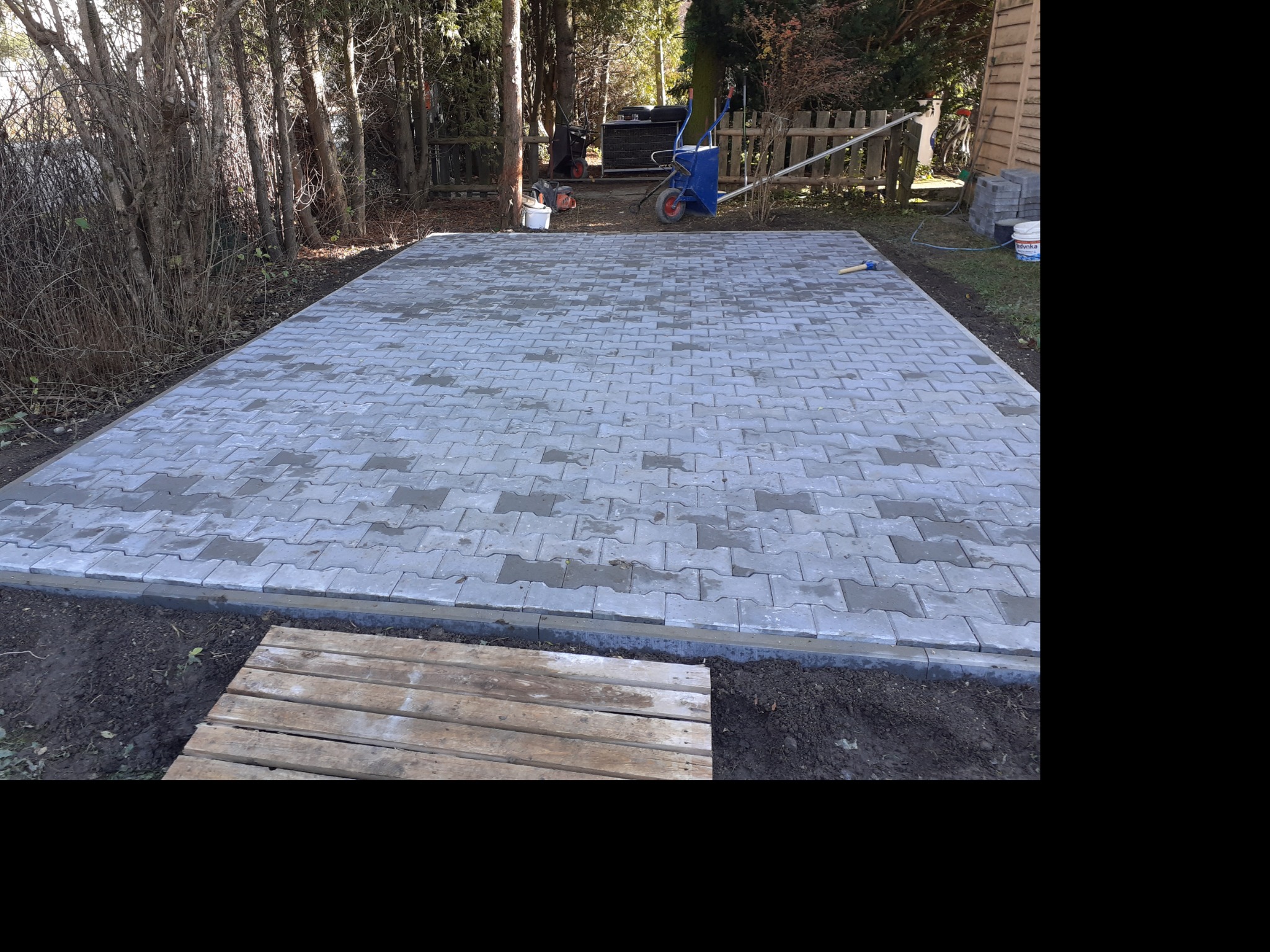 Nowo ułożona szara kostka brukowa w ogrodzie, widoczna paleta drewniana, narzędzia i taczka w tle, częściowo w cieniu drzew.