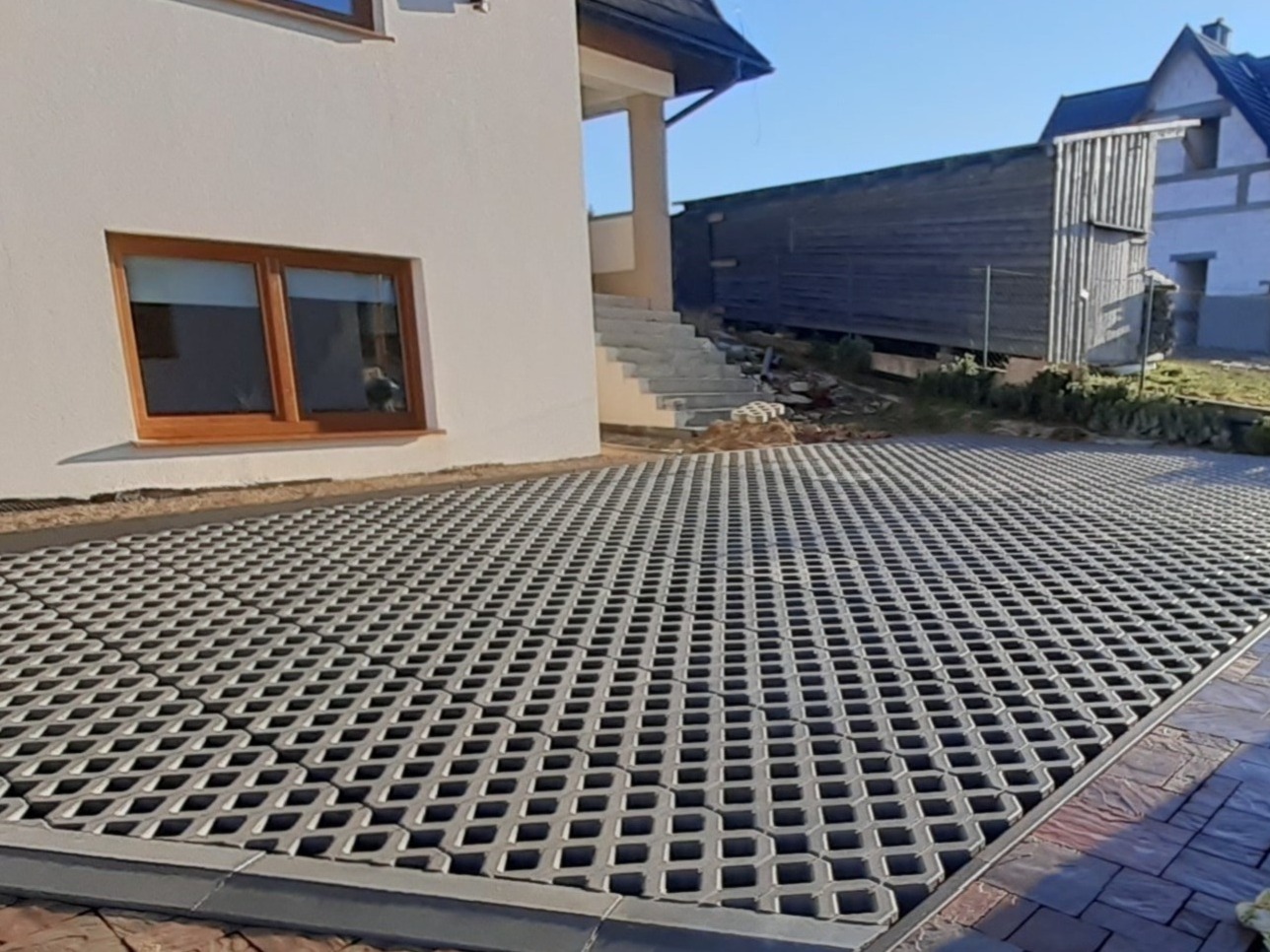 Ułożone ażurowe płyty betonowe na podjeździe przed domem, widoczne schody wejściowe i fragment elewacji z oknem.