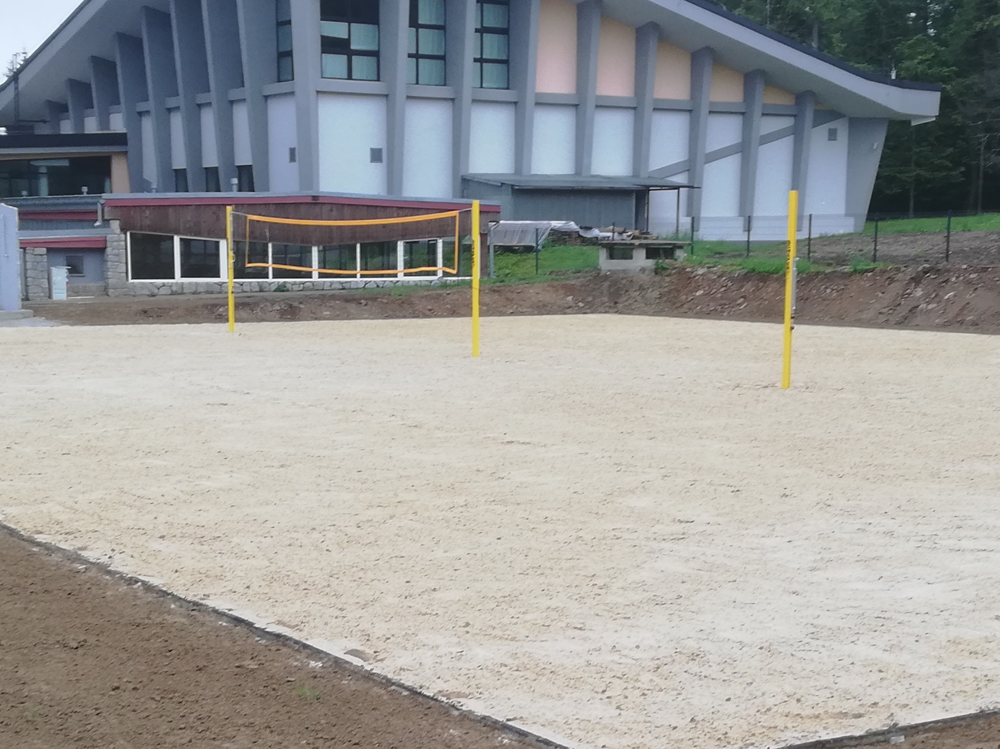Przygotowany teren pod boisko do siatkówki plażowej z żółtą siatką i słupkami, obok budynku z dużymi oknami i terenem z nasadzeniami.