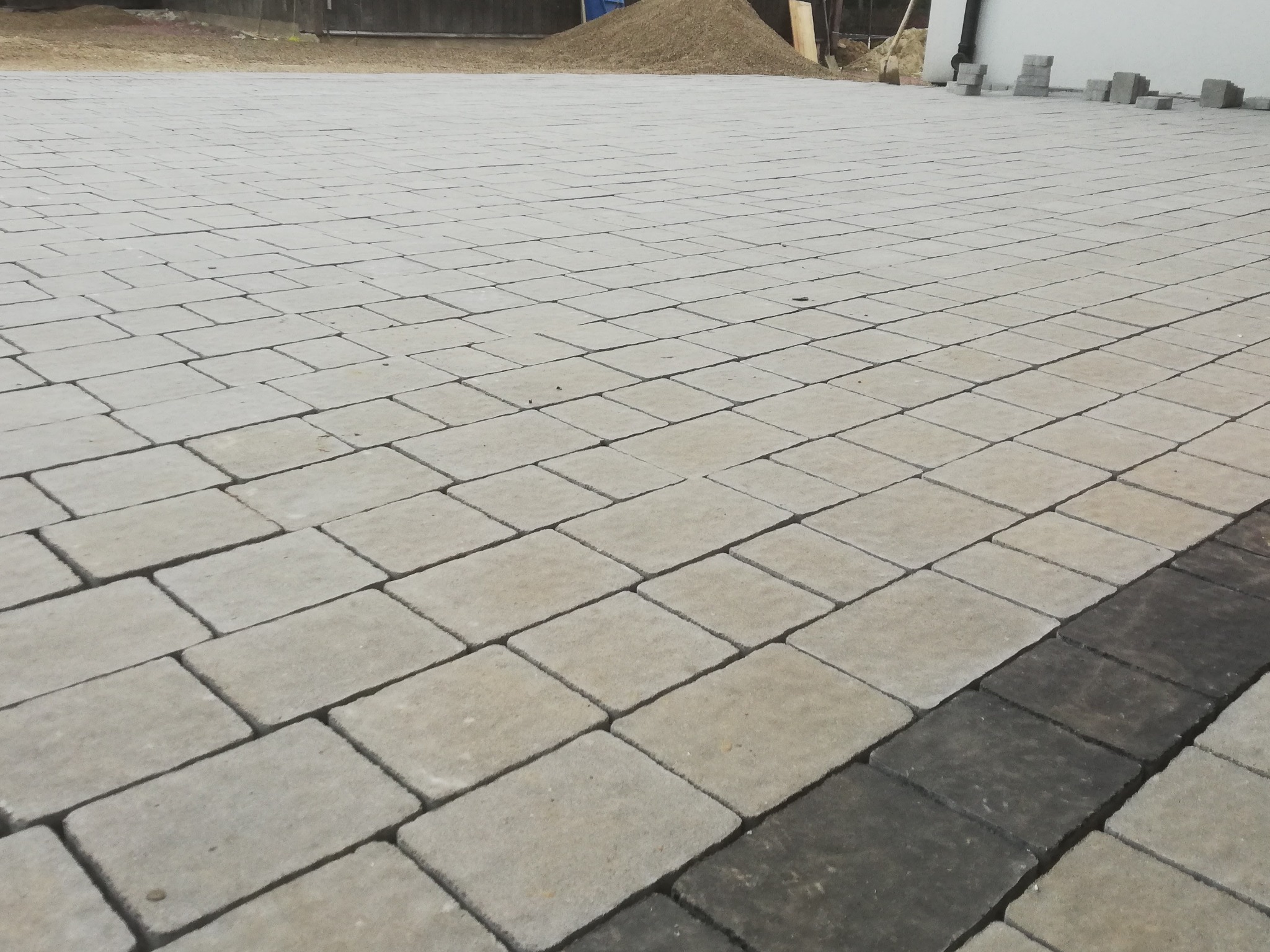Ułożona jasnoszara kostka brukowa z ciemniejszym pasem obrzeża, widoczny stos kostki i narzędzia w tle.