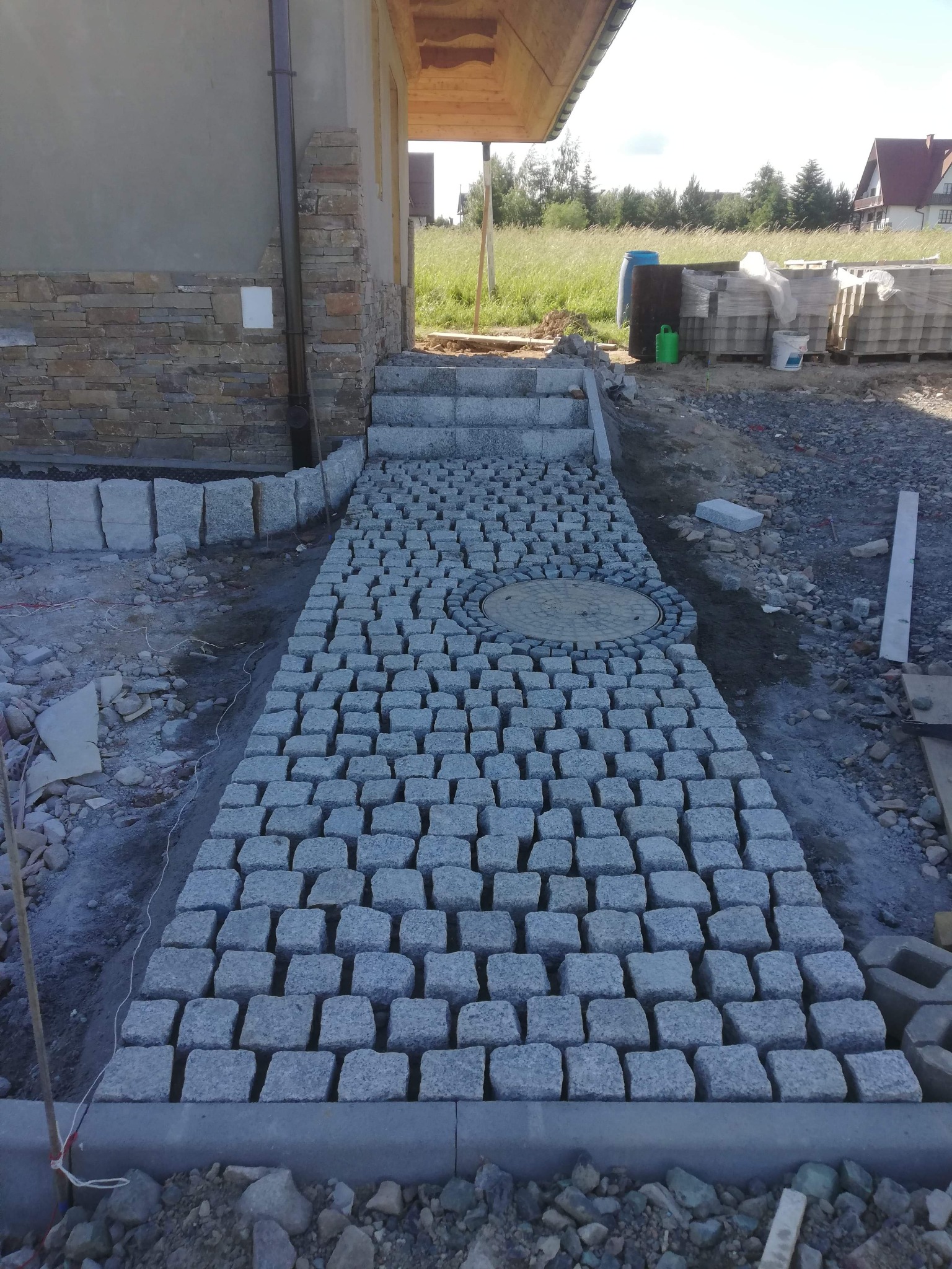 Ścieżka z szarej kostki granitowej prowadząca do domu, z okrągłym włazem wkomponowanym w nawierzchnię, widoczne materiały budowlane w tle.