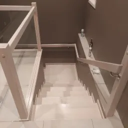 Widok z góry na jasne, drewniane schody z przeszkloną balustradą, prowadzące w dół. Na półce przy ścianie widoczne akcesoria montażowe i preparaty czyszczące.