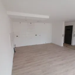 Wnętrze w trakcie prac: częściowo położone płytki na ścianie w kuchni, widoczne puszki elektryczne, surowa ściana przygotowana pod zabudowę, jasna podłoga imitująca drewno, czarne drzwi w tle.