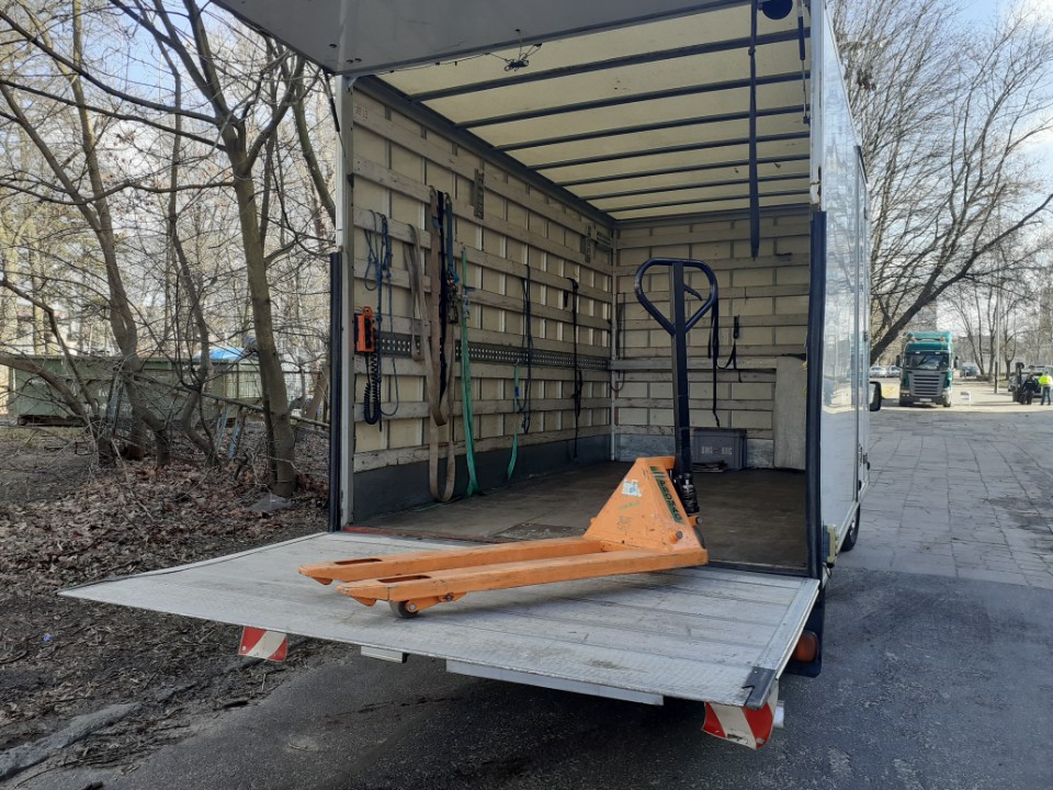 Wnętrze otwartego busa transportowego z wózkiem paletowym na rampie załadunkowej, widoczne pasy mocujące na ścianie i drzewa w tle.