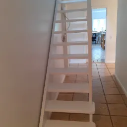 Białe, strome schody wewnątrz domu, prowadzące na poddasze lub wyższą kondygnację, z widocznym kablem elektrycznym i wnętrzem pokoju w tle.