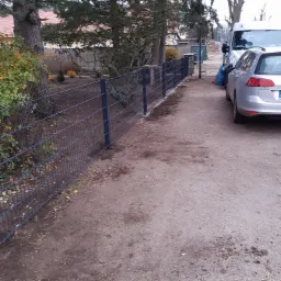 Zamontowane ogrodzenie panelowe z ciemnego drutu na posesji, widoczne drzewa i zaparkowane samochody w tle.