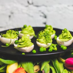 Po Pierwsze Spółka z o.o. - Wielkanocne jajka faszerowane zielonym kremem z groszkiem, podane na czarnym talerzu w otoczeniu tulipanów.