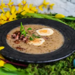 Żurek wielkanocny w czarnym talerzu, udekorowany jajkiem na twardo, gałązkami i bazią, na mchu z pisankami i żółtymi kwiatami forsycji.