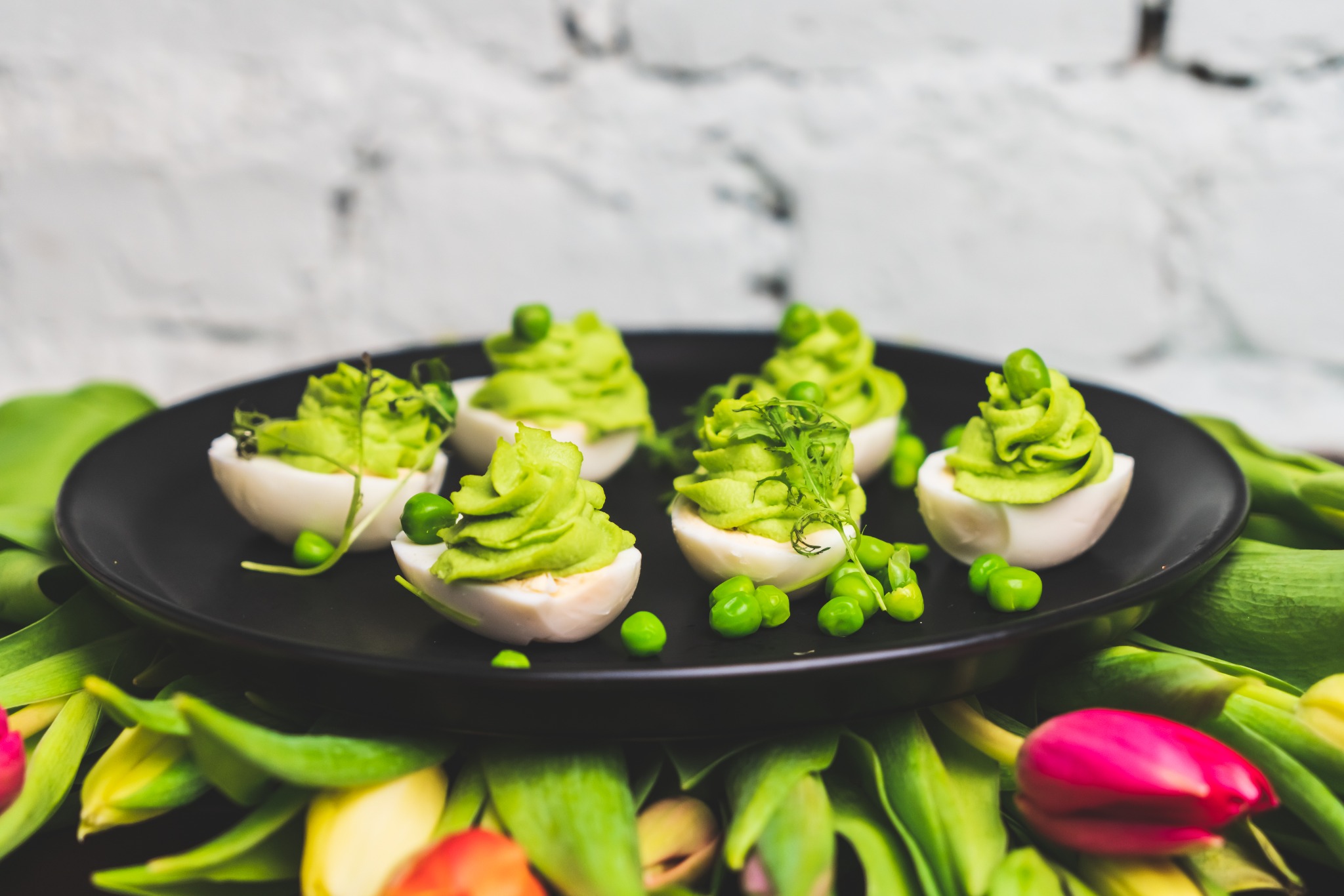 Wielkanocne jajka faszerowane zielonym kremem z groszkiem, podane na czarnym talerzu w otoczeniu tulipanów.