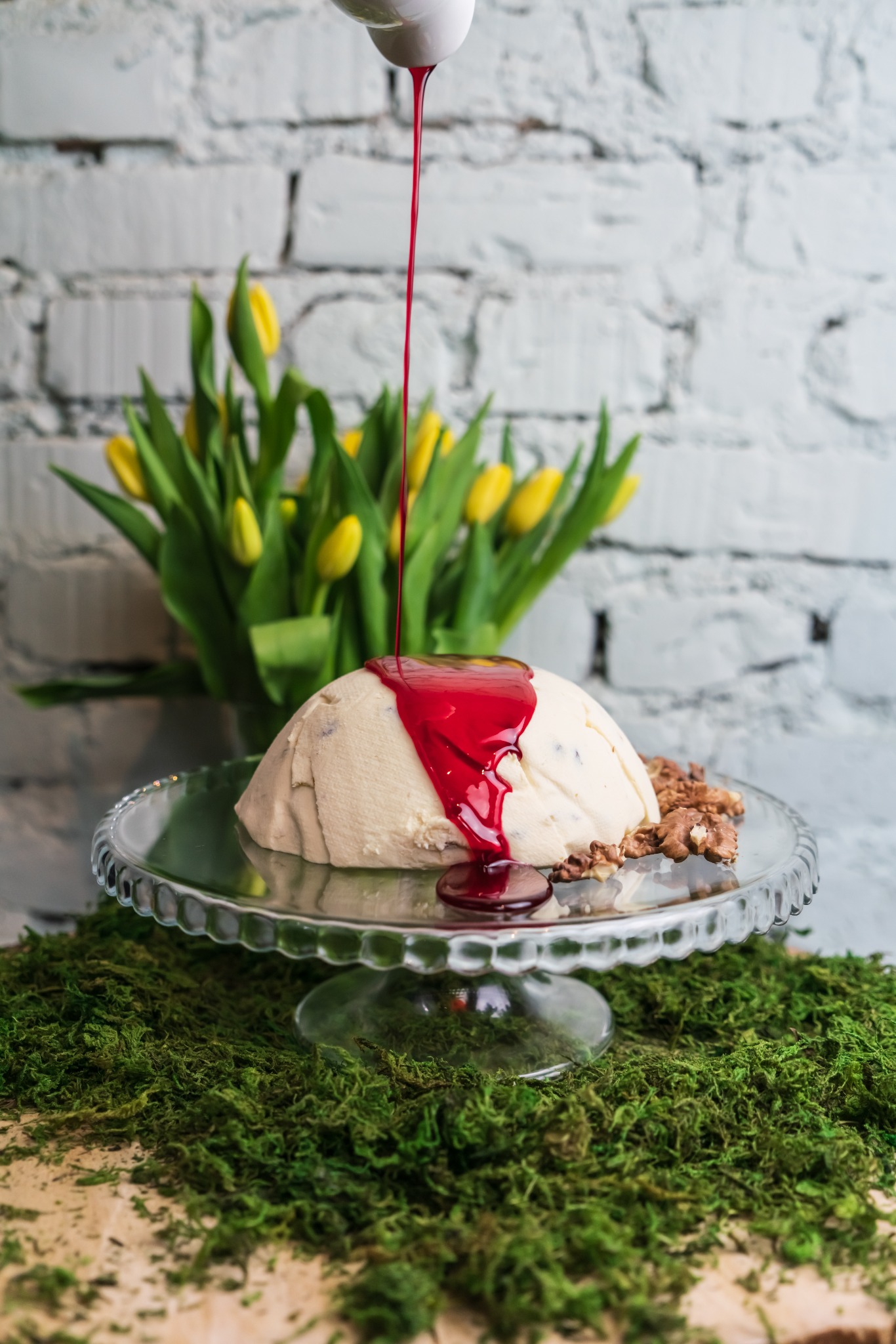 Pascha wielkanocna na szklanym paterze, polewana sosem z czerwonych owoców, obok orzechy włoskie, w tle bukiet żółtych tulipanów i biała ściana z cegły.