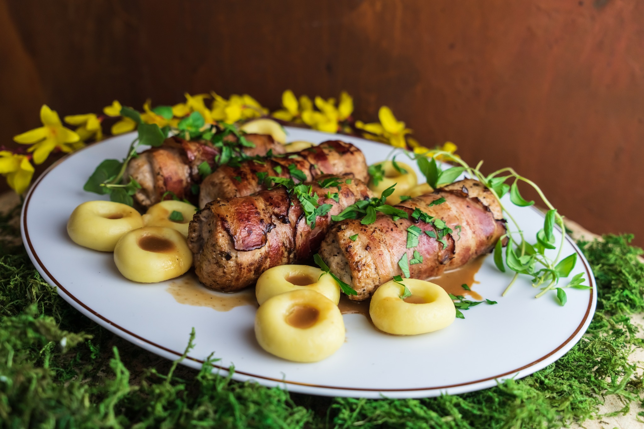 Roladki z mięsa owinięte boczkiem, udekorowane świeżymi ziołami i podane z kluskami śląskimi na białym talerzu z brązową obwódką, w tle żółte kwiaty i brązowa ściana.
