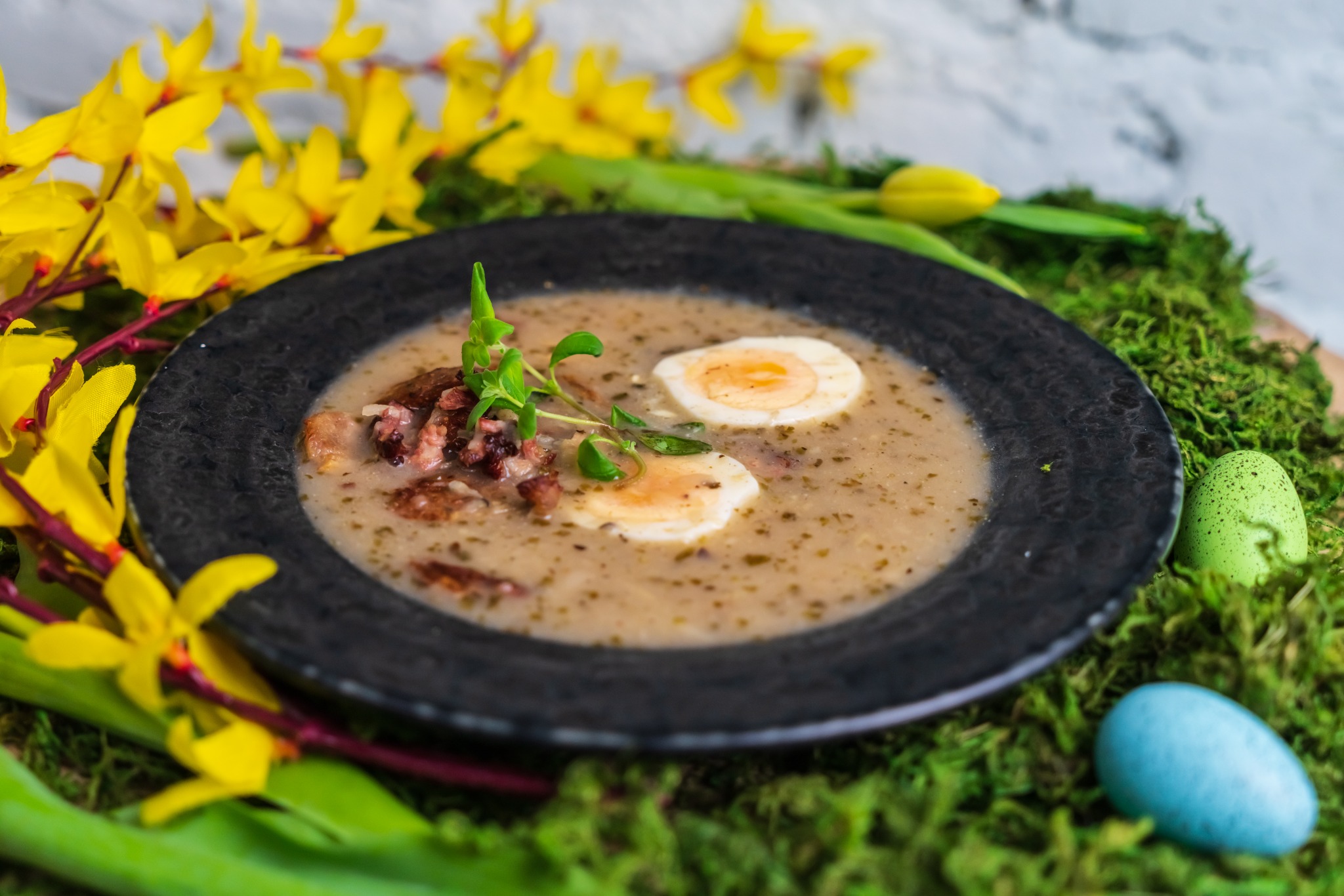 Żurek wielkanocny w czarnym talerzu, udekorowany jajkiem na twardo, gałązkami i bazią, na mchu z pisankami i żółtymi kwiatami forsycji.