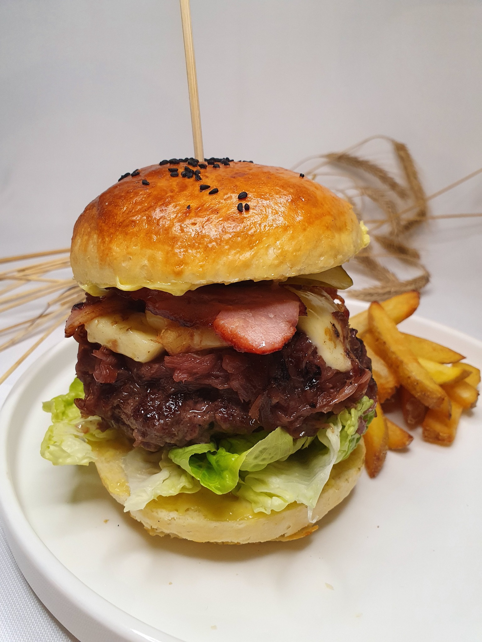 Apetyczny burger z wołowiną, karmelizowaną cebulą, boczkiem i serem, podany z frytkami na białym talerzu, dekorowany kłosami zbóż w tle.