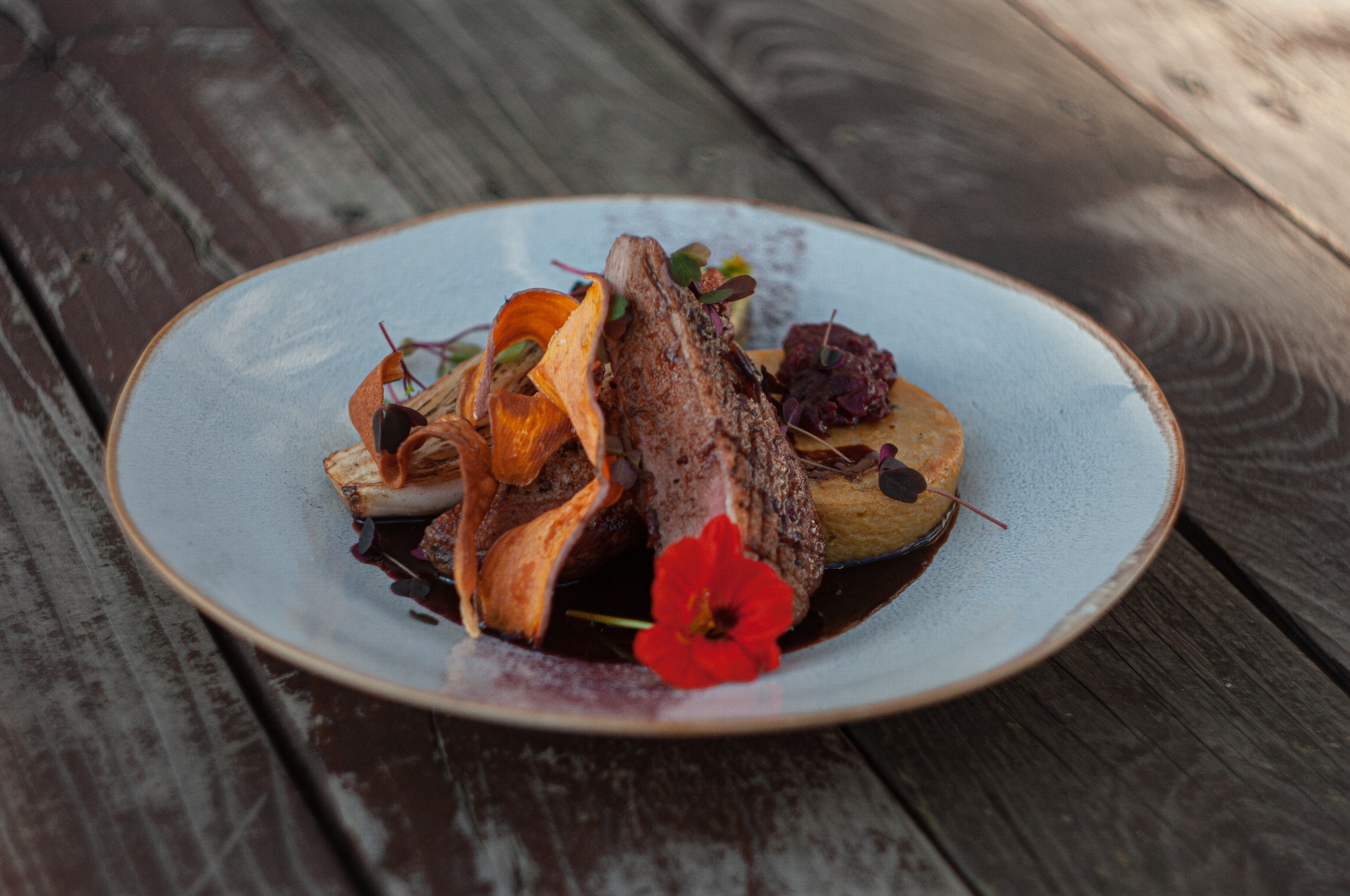 Elegancko podane danie mięsne z puree, chipsami z batatów i czerwoną nasturcją na talerzu z szarobłękitną obwódką, ułożone na blacie z surowego drewna.