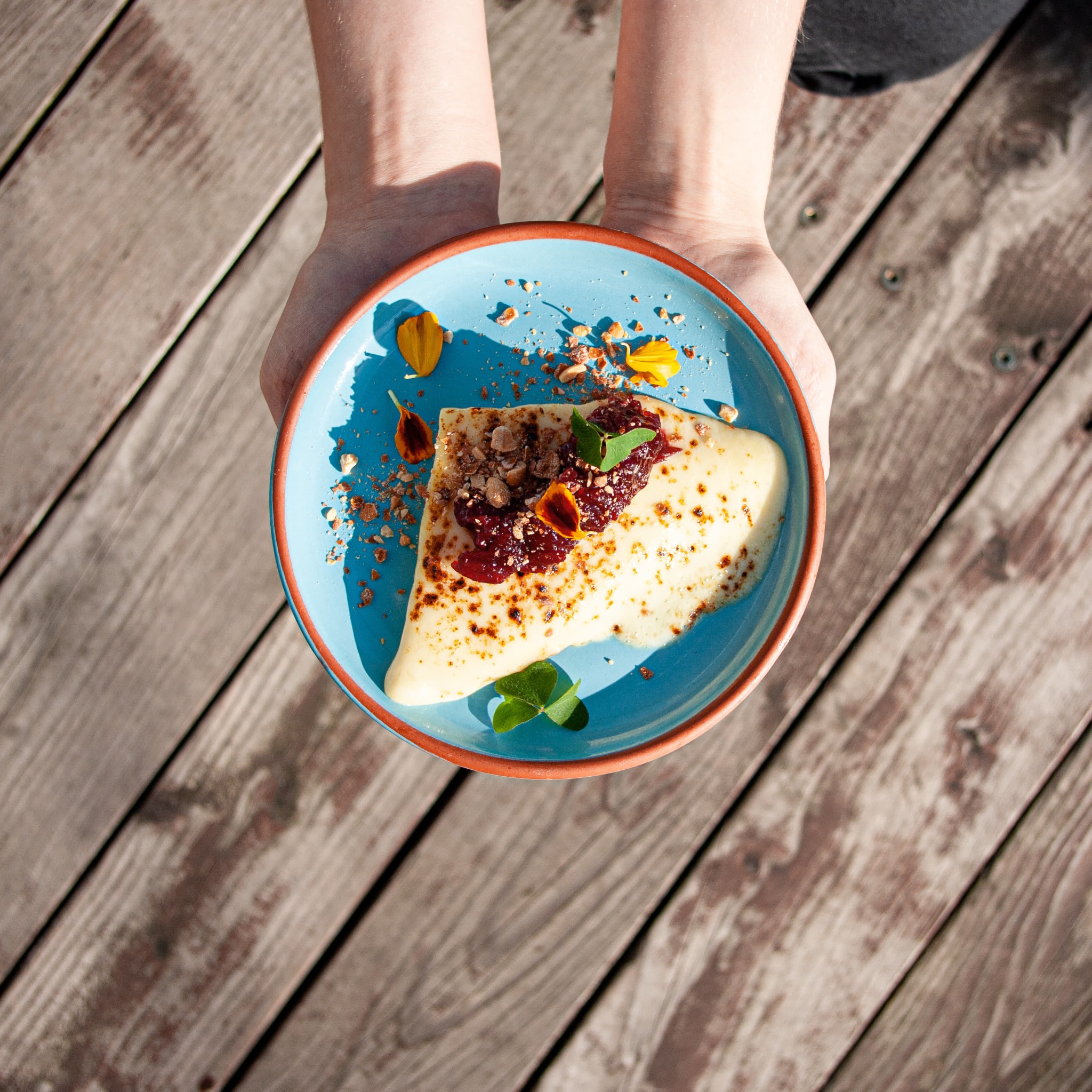 Naleśnik z dżemem i posypką orzechową, udekorowany kwiatami i koniczyną, podawany na turkusowym talerzu z pomarańczowym brzegiem, trzymany przez dwie ręce na tle drewnianych desek.