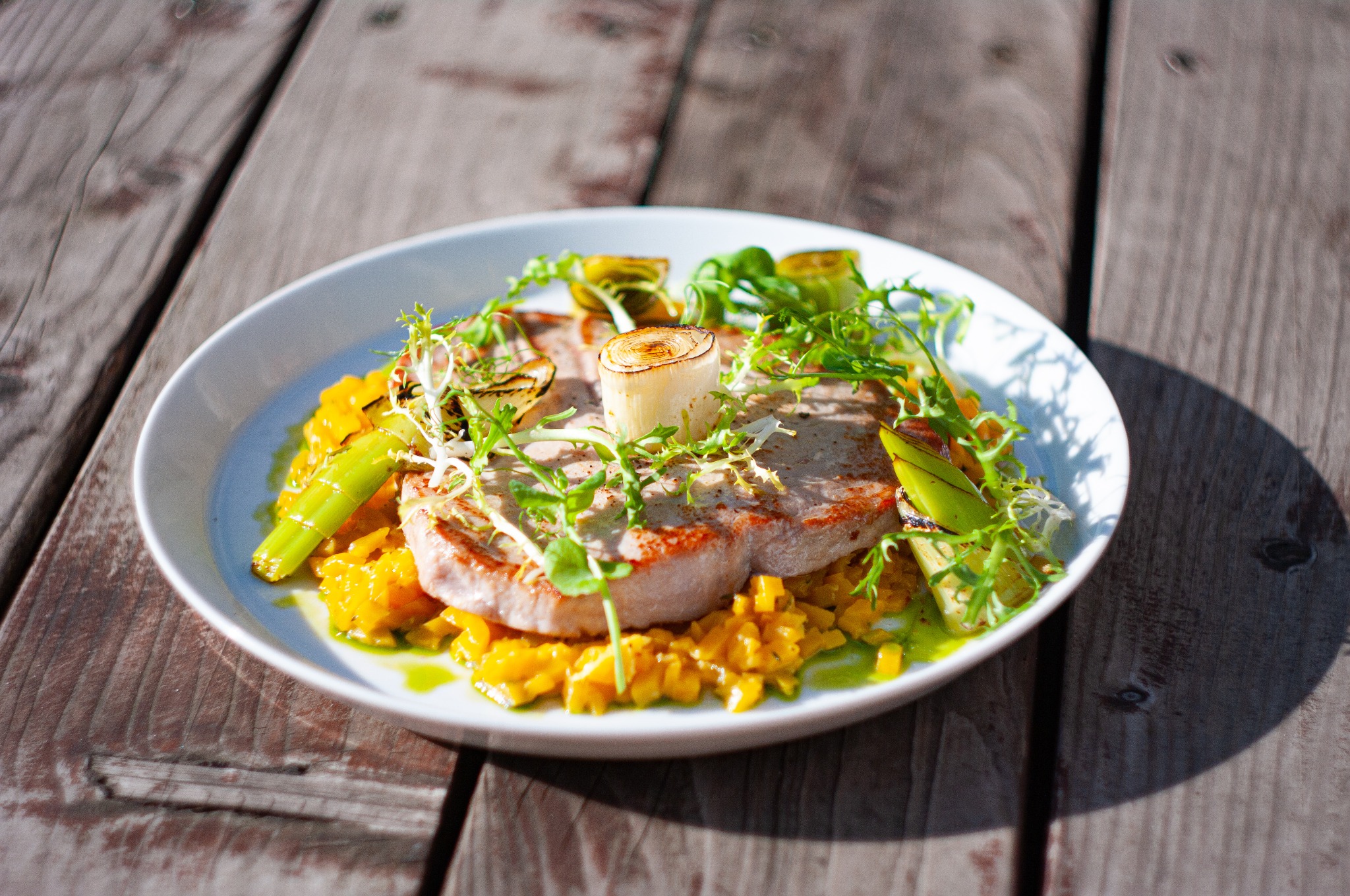 Elegancko podane danie z grillowaną rybą, żółtym risotto i warzywami na białym talerzu, umieszczone na drewnianym stole w plenerze.