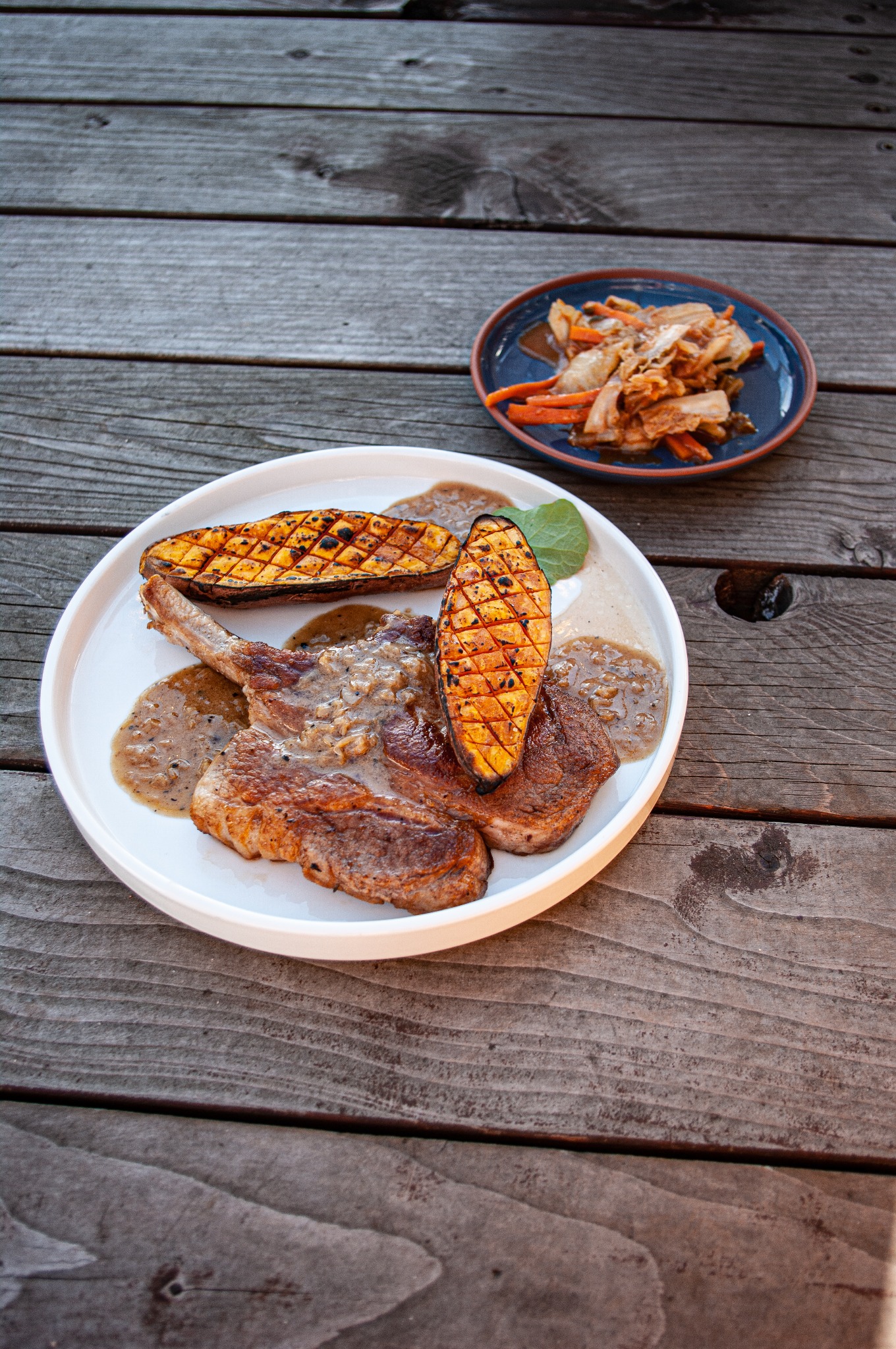 Dwa kotlety wieprzowe z kością, polane sosem pieprzowym, podane z grillowaną dynią piżmową z kratką i listkiem mięty na białym talerzu, obok sałatka z kapusty i marchewki na niebieskim talerzu,...