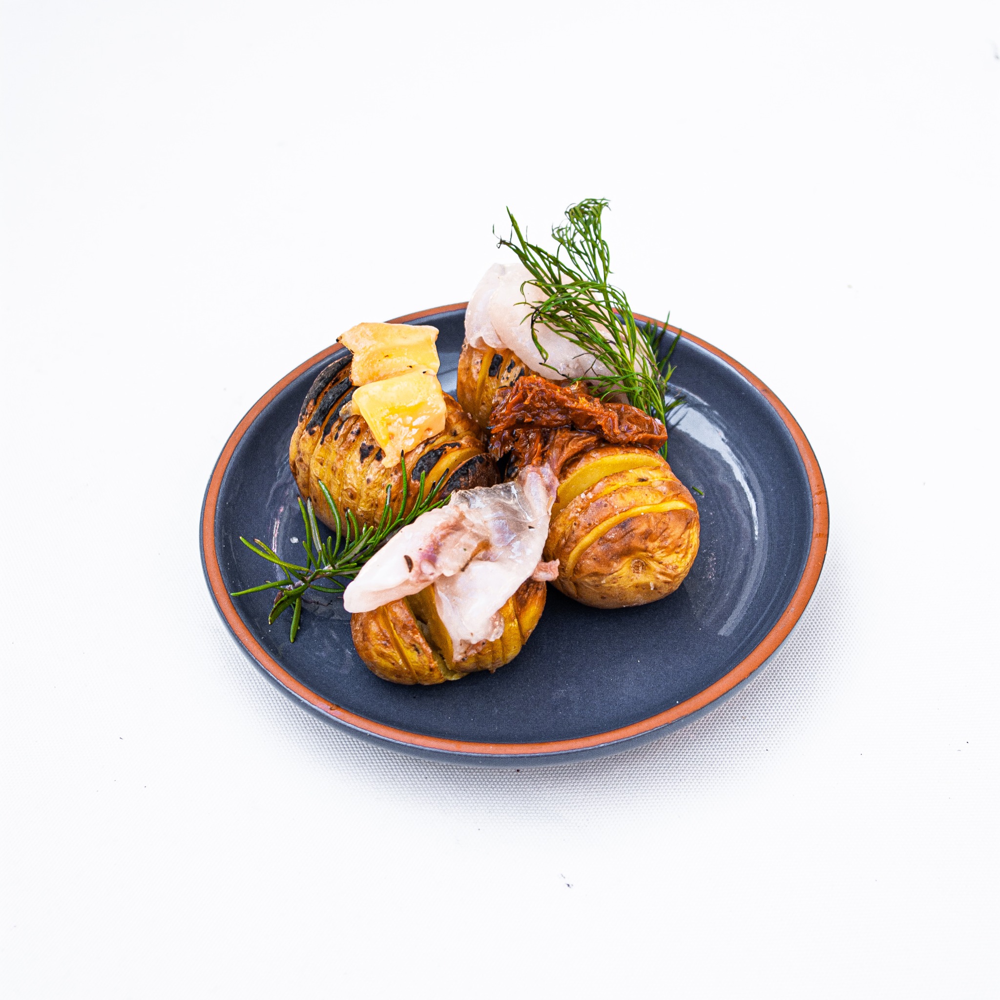 Elegancko podane pieczone ziemniaki hasselback z dodatkami: ryba, suszone pomidory, ser i zioła na ciemnym talerzu z pomarańczowym brzegiem, na białym tle.