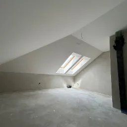 Poddasze z dwoma oknami dachowymi, ściany przygotowane pod malowanie, widoczny przewód elektryczny i odkurzacz przemysłowy.