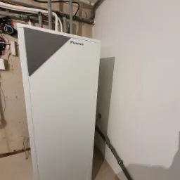 Biała pompa ciepła Daikin z szarym panelem na tle surowej ściany z rurami instalacji grzewczej i fragmentem białej ściany, widok z boku.