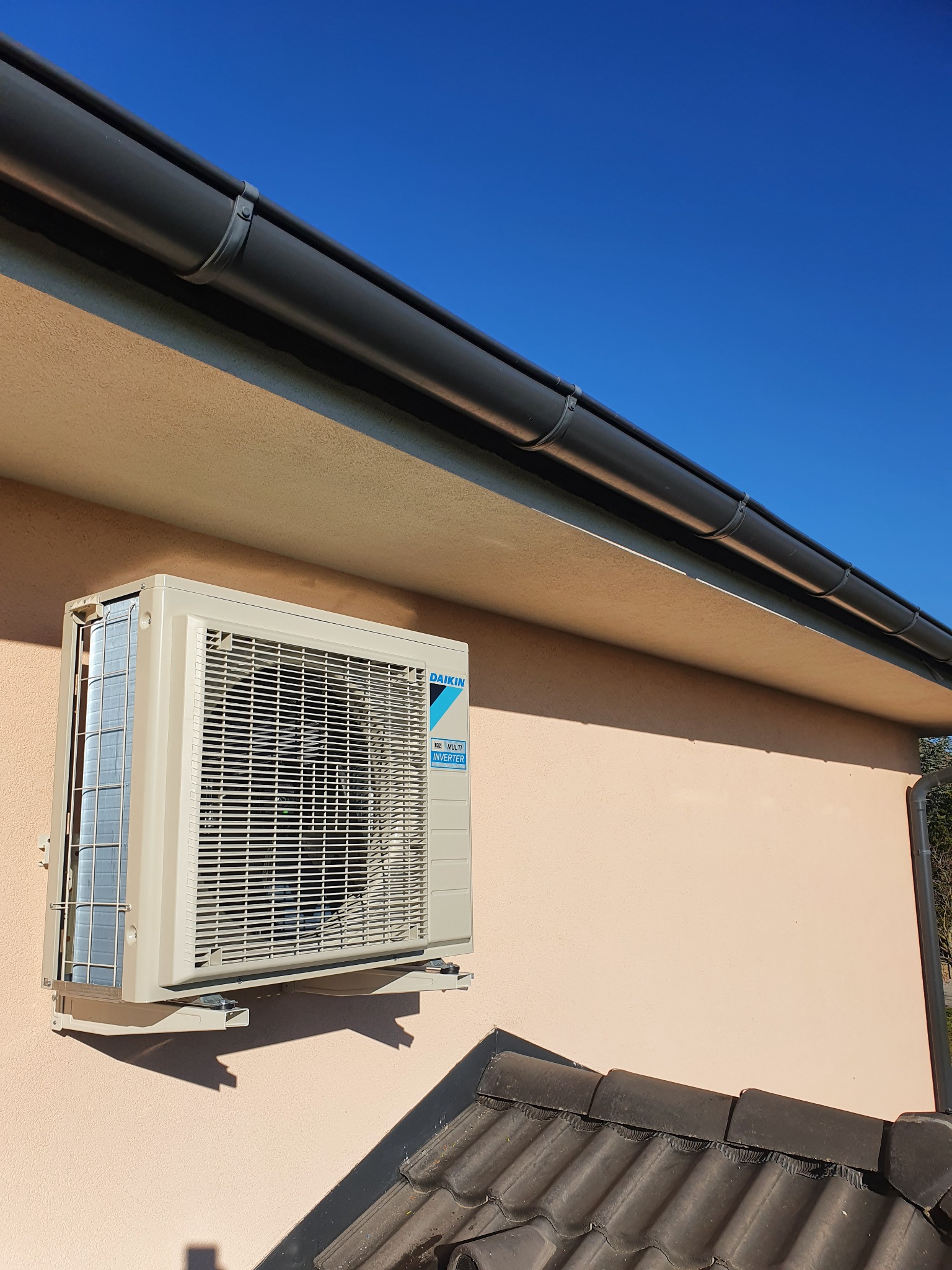 Zewnętrzna jednostka klimatyzacji Daikin zamontowana na elewacji budynku z widocznym dachem i błękitnym niebem w tle.