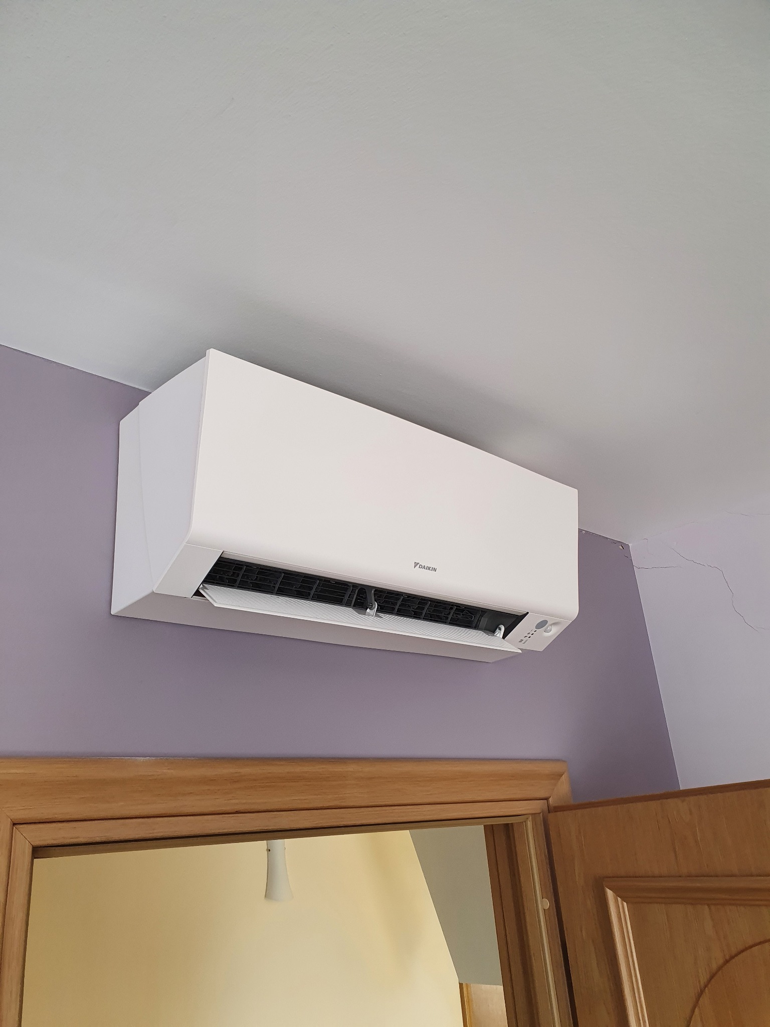 Biały klimatyzator marki Daikin zamontowany na fioletowej ścianie nad drewnianą framugą drzwiową, widoczne pęknięcie na ścianie w górnym rogu.